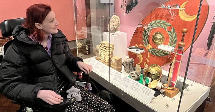 Kate Stanforth sat in wheelchair looking at exhibition inside The Museum of Archaeology, Palace Green Library